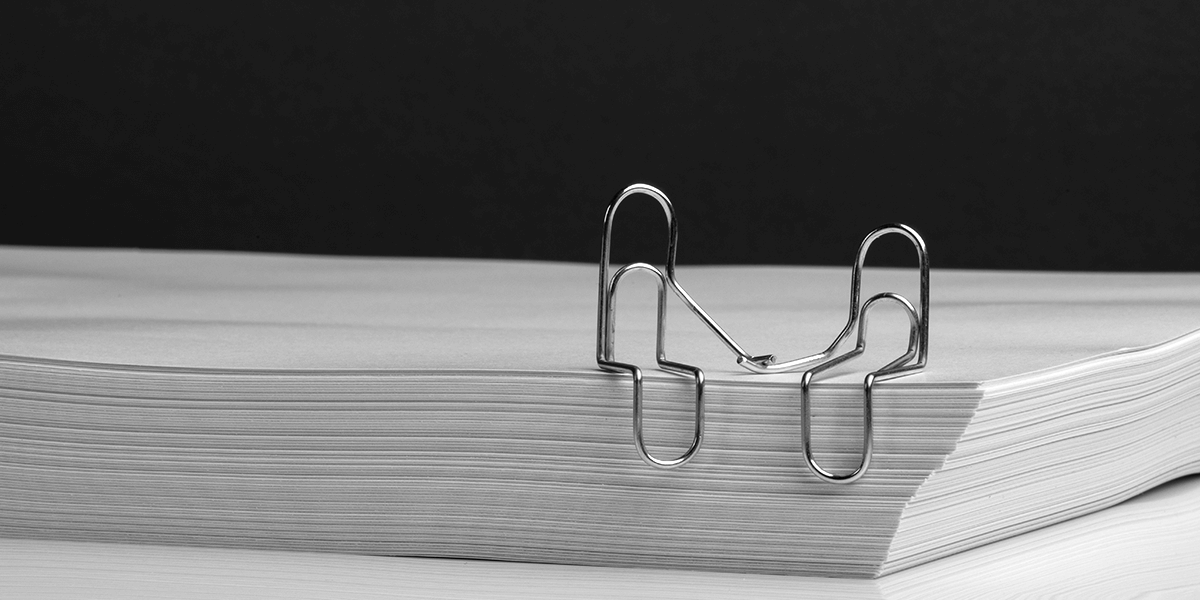 Two paperclicks set on the edge of a stack of papers, the paperclips are bent to resemble two people sitting beside each other holding hands.