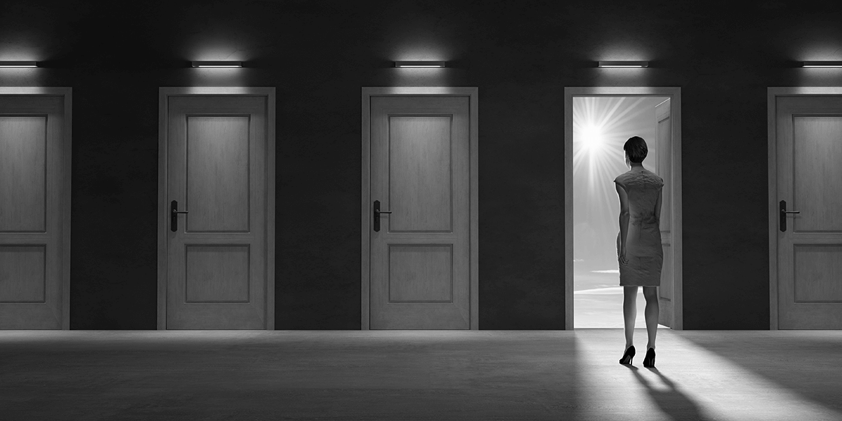 Dark hallway of closed doors, one door is open and a woman is entering the room.
